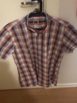 Rutig kortärmad skjorta Louis Barton - Snygg kortärmad skjorta från Louis Barton i regular fit. Skjortan har ett rutigt mönster i rött, blått, beige och brunt, med klassisk krage och knappar framtill. Perfekt för dig som gillar färg och vill ha en chill look.