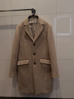 Beige kappa från These Glory Days - Snygg beige kappa med klassisk krage och tre knappar framtill. Jackan har två stora fickor med lock och är helfodrad med ljus insida. Perfekt för dig som gillar stilrena och tidlösa plagg med en modern touch. Jag som haft den är 183cm. Nypris 2500kr mitt pris 500kr