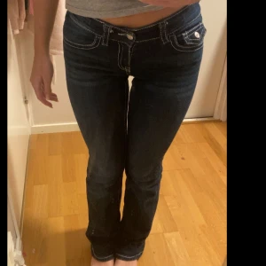 Mörkblå bootcut jeans Gina Tricot - Säljer ett par mörkblå jeans från Gina Tricot med bootcut passform och vita kontrastsömmar. Jeansen har låg midja, dekorativa fickor med lock och knapp bak, dessa jeans är slutsålda och dem är i storlek 158