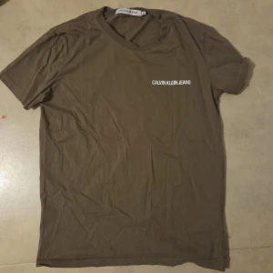 Brun t-shirt från Calvin Klein Jeans - Säljer en brun t-shirt från Calvin Klein Jeans i storlek XL/L/M. T-shirten har korta ärmar och en enkel, clean design med vit logotyp på bröstet. Tillverkad i 100% bomull för skön och mjuk känsla. Perfekt för dig som gillar stilrena och klassiska plagg. Inget fel på tröjan den är bara skrynklig, se till att vända ut och in på tröjan när den ska tvättas