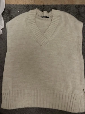 Beige stickad väst från Bik Bok - Säljer en beige stickad väst från Bik Bok i storlek S. Västen har en bred ribbad v-ringad krage och ribbade detaljer vid nederkanten. Perfekt att styla över en skjorta eller t-shirt för en chill look.