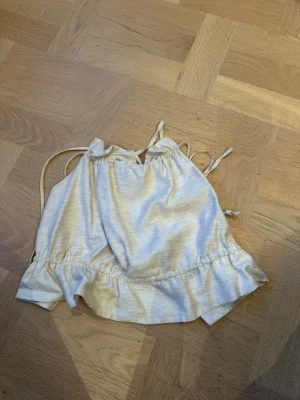 Ljusbeige linnetopp från Gina Tricot - Supersöt ljusbeige linnetopp från Gina Tricot med tunna axelband och knyt i sidorna. Toppen har en avslappnad passform och lätt glansigt tyg som känns mjukt mot huden. 