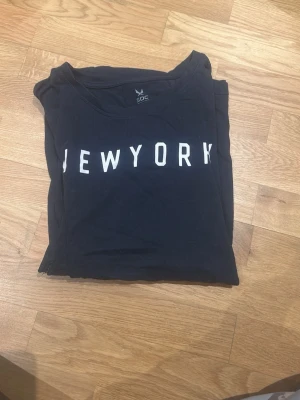 Svart t-shirt SOC med New York-tryck - Svart t-shirt från SOC i storlek M med vit text 'NEW YORK' på bröstet. Klassisk rund hals och korta ärmar. Materialet är mjuk bomull som känns skönt mot huden. Perfekt för dig som gillar enkel streetstyle och vill ha en chill vibe.