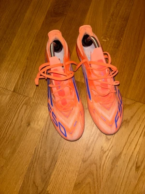 Adidas F50 orange fotbollsskor - Säljer ett par Adidas F50 fotbollsskor i en riktigt fet orange färg med blå detaljer och snörning. Skorna har en spetsig tå och platt sula med dobbar för gräsplan. Lätta och smidiga med syntetmaterial som ger bra känsla på bollen. Perfekta för dig som vill sticka ut på planen.