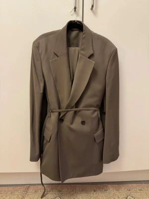 Beige kavaj med bälte från Acne Studios - Säljer en snygg beige kavaj från Acne Studios med bälte i midjan och dubbelknäppning. Kavajen har klassisk krage, långa ärmar och fickor framtill. Perfekt oversized passform för en trendig look.