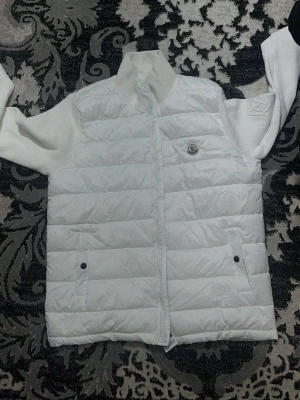 Vit pufferjacka från Moncler - Säljer en fräsch vit pufferjacka från Moncler med stickade ärmar och hög krage. Jackan har dragkedja framtill, två fickor med knapp och ett broderat Moncler-märke på bröstet. Perfekt för dig som gillar stilren och clean look.