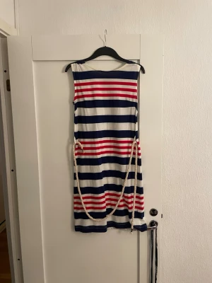 Randig sommarklänning från Ellos - Säljer en ärmlös klänning från Ellos med breda horisontella ränder i marinblått, rött och vitt. Klänningen har en kort längd och ett rep som bälte i midjan för extra detalj. Perfekt för varma dagar och har en mjuk bomullskänsla.