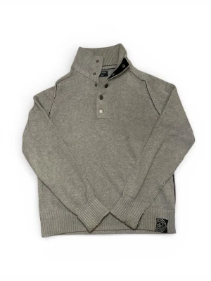 Grå stickad tröja från East West - Snygg grå stickad tröja från East West i storlek medium. Tröjan har hög krage med flera metallknappar framtill och ribbade muddar vid ärmslut och nederkant. Tröjan har sjukt bra kvalitet och är väldigt varm, perfekt för en kall vinterdag.