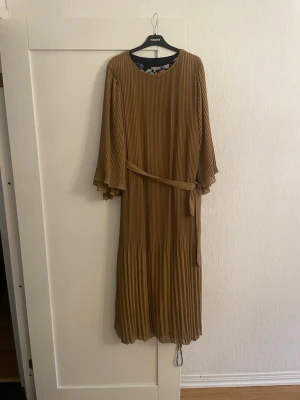 Plisserad långklänning med bälte - Säljer en snygg plisserad långklänning i brun ton från KappAhl. Klänningen har rund hals, vida långärmar och ett matchande bälte i midjan. Materialet känns lätt och luftigt, perfekt för dig som gillar stilrena och bekväma plagg.