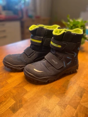 Svarta vinterstövlar från Superfit - Säljer ett par mörktblåa vinterstövlar från Superfit med grönt fleecefoder och blå detaljer. Skorna har grov sula för extra grepp, två kardborreband och är tillverkade i syntetmaterial och mocka. Perfekta för kalla dagar och snöiga äventyr.  https://www.zalando.se/superfit-husky-vinterstoevlar-blaugruen-le314k01q-k12.html