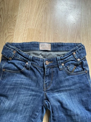 Vintage jeans - Säljer ett par blå ginatricot jeans med bootcut passform och klassisk femficksdesign. Jeansen har snygga detaljer som bruna nitar, dekorativa sömmar och ett justerbart spänne bak i midjan. Materialet är slitstarkt jeans och färgen är mörkblå med lätt tvättad look.