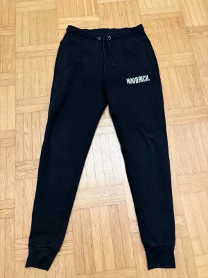 Svarta mjukisbyxor från Hoodrich XS - Svarta mjukisbyxor från Hoodrich i storlek XS. Byxorna har snörning i midjan, ribbade muddar vid bensluten och tryckt Hoodrich-logga på ena benet. Tillverkade i mjukt bomullsmaterial, perfekta för chill eller träning.