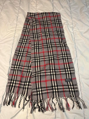 Grå rutig Burberrys halsduk - Säljer en klassisk grå halsduk från Burberrys med ikoniskt rutigt mönster i svart, vitt och rött. Handduken är tillverkad i 100% lammull och har snygga fransar längs nederkanten. Perfekt för dig som gillar tidlös design och vill sticka ut med ett lyxigt plagg. N19