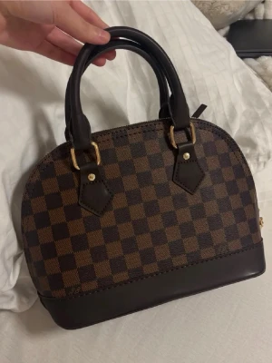 Alma BB väska - BUDA RIMLIGT! Säljer en snygg handväska med klassiskt brunt och mörkbrunt rutigt Damier-mönster. Väskan har guldfärgade detaljer, lås och dragkedja, samt mörkbruna handtag i skinn. Insidan är klarröd och har en innerficka med dragkedja. Använd en gång och i otroligt bra skick.❤️