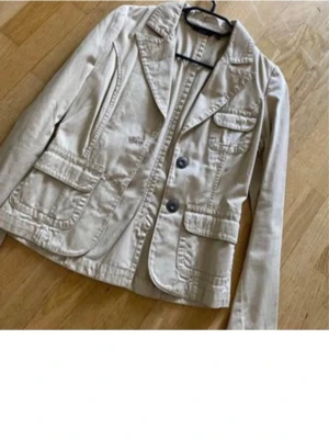 Beige kavaj från Gina XS - Snygg beige kavaj från Gina i storlek XS. Jackan har klassisk krage, två svarta knappar framtill och flera fickor med lock. Materialet är bomull och passformen är normal. Perfekt för dig som gillar stilrena och enkla plagg. Fint skick  fri frakt 