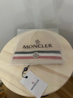 Beige Moncler mössa med ränder - Säljer en helt ny mössa oanvänd, Storlek One size Fraktar snabbt inom 24h efter köp.Ställ gärna din fråga.