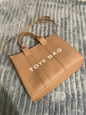 Beige tote bag med axelrem - Säljer en beige tote bag med tydlig text på framsidan. Väskan har både handtag och en justerbar axelrem i samma färg. Tillverkad i skinnimitation med stilrena detaljer i guld. Perfekt storlek för att få plats med allt du behöver under dagen.