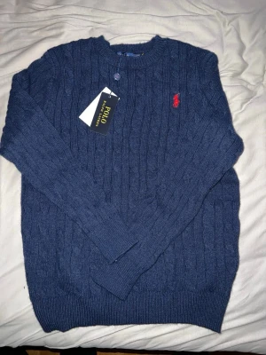 Marinblå stickad tröja Polo Ralph Lauren - Snygg marinblå stickad tröja från Polo Ralph Lauren i storlek S. Tröjan har kabelstickat mönster, rund hals och en röd broderad logga på bröstet. Perfekt för dig som gillar klassisk stil med en modern touch.