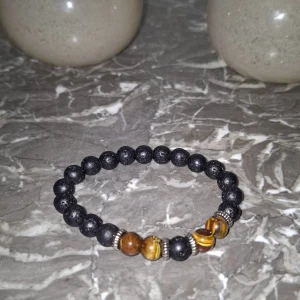 Svart och brun pärlarmband med stenar - Stilrent armband med svarta lavapärlor och bruna tigeröga-stenar, separerade av små silvriga detaljer. Elastiskt band gör det enkelt att ta på och av. Perfekt accessoar för att lyfta din outfit med en cool och jordnära vibe.