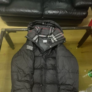 Svart Burberry pufferjacka M - Svart pufferjacka från Burberry med klassisk rutig huva i grått, vitt och rött. Jackan har quiltad design, dragkedja och knappar framtill samt en patch med Burberry London England på axeln. Perfekt för kalla dagar med snygg streetstil.