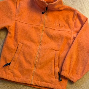 Orange fleece zip-up - Säljer en orange fleecejacka från James & Nicholson med dragkedja framtill och två sidofickor. Jackan har hög krage och elastiska muddar vid ärmslut. Perfekt för kyliga dagar och enkel att slänga på över en hoodie. Storlek: 122/128