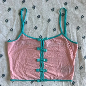 Rosa linnetopp med turkosa rosetter - Säljer en söt rosa linnetopp från Dolls Kill x Hatsune Miku med turkosa kantband och justerbara axelband. Framtill sitter fem turkosa rosetter som ger en gullig vibe. Toppen är croppad och gjord i mjuk bomull, perfekt för dig som gillar färgglada detaljer. Använt en eller två gånger. Måtten finns vid behöv. 