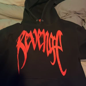 Svart Revenge hoodie med röd print - Svart hoodie från Revenge med stor röd logga framtill, fet röd handavtrycksprint på ryggen och texten 'Revenge Is In The Creators Hands' på ärmen. Klassisk huva och känguruficka, tillverkad i mjuk bomullsmix. Perfekt för dig som gillar streetwear med attityd.