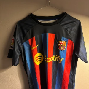 FC Barcelona Lewandowski matchtröja - Säljer en officiell FC Barcelona matchtröja från Nike med Lewandowski och nummer 9 på ryggen. Tröjan har klassiska blå och röda ränder, gula detaljer och klubbmärke på bröstet. Materialet är lätt och andas, perfekt för fotboll eller supporterliv.