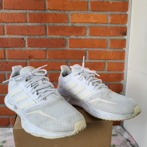 Vita Adidas sneakers strl 42 - Fräscha vita Adidas sneakers med mesh-ovandel och klassiska tre ränder på sidan. Skorna har snörning och platt sula, perfekt för dig som gillar sportig stil. Lätta och ventilerande med syntetdetaljer och rund tå. Snygga att matcha med jeans eller shorts.
