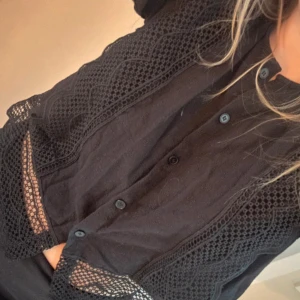 Svart kortärmad blus med spetsdetaljer - Svart blus med kort ärm och snygga spets- och broderidetaljer. Knappar framtill och coola meshpartier längs axlar och nederkant. Skjortan har en avslappnad passform och är perfekt för dig som gillar unika detaljer. Storlek M men passar en S också.