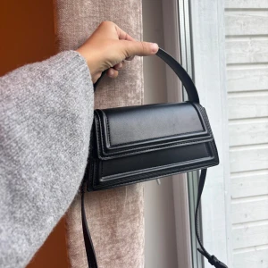 Svart stilren axelväska i skinn från Zara - Snygg svart axelväska i slätt skinn med rektangulär form och stilrena detaljer. Väskan har ett brett handtag och justerbar axelrem med silvriga metalldetaljer. 