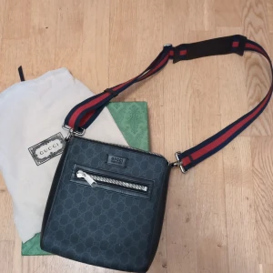 Svart axelväska från Gucci bra skick bara en liten skråma 8/10 - Snygg svart axelväska från Gucci med klassiskt GG-mönster och bred justerbar axelrem i rött och marinblått. Väskan har en dragkedjeficka framtill och detaljer i skinn. Perfekt för dig som vill ha en stilren och lyxig accessoar.