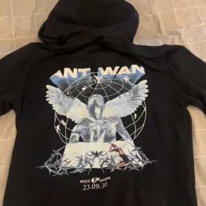 Svart Ant Wan hoodie från Dopest - Svart hoodie från Dopest x Ant Wan med stor grafisk print på ryggen och mindre tryck på bröstet. Tröjan har huva med snörning och ribbade muddar. Materialet är mjuk bomull, perfekt för dig som gillar streetwear och snygga detaljer.