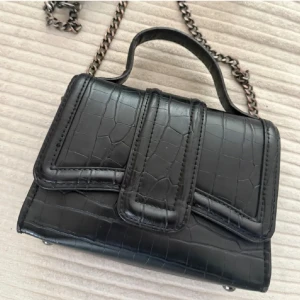 Svart axelväska med kedja frå Zara  - Snygg svart axelväska med croco-mönstrad yta och cool kedjerem. Väskan har ett stilrent och modernt utseende med både handtag och axelrem, perfekt för att lyfta din outfit. Materialet är skinnimitation och designen är kompakt men rymlig för det viktigaste.