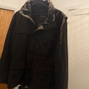 Mörkbrun parkajacka från MooRER - Parkajacka från MooRER i mörkbrunt med flera stora fickor framtill och hög krage. Jackan har dragkedja och knappstängning, samt en loose passform. Materialet är slitstark polyester och jackan är långärmad, perfekt för kyliga dagar.