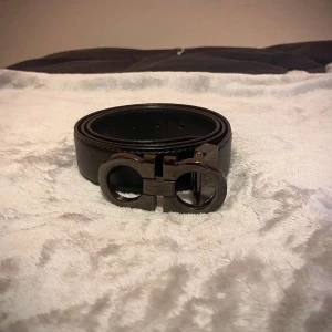 Svart Ferragamo skärp med metallspänne - Snyggt svart skärp från Ferragamo med ett ikoniskt, rundat metallspänne i mörk silverfärg. Bältet är tillverkat i slätt skinn och har stilrena sömmar längs kanterna. Perfekt för dig som vill ha en lyxig och modern accessoar med tydlig designerstil.