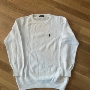 Vit stickad tröja från Ralph Lauren - Snygg vit stickad tröja från Ralph Lauren med klassisk rund hals och broderad logga på bröstet. Tröjan har långärmad passform och ribbade muddar vid ärmslut och nederkant. Perfekt för dig som gillar stilrena och tidlösa plagg.
