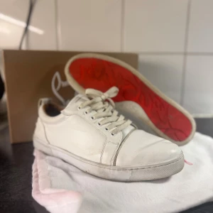 Vita sneakers från Christian Louboutin - Säljer ett par vita sneakers från Christian Louboutin med ikonisk röd sula och klassisk snörning. Skorna är i skinn och har en stilren design med rund tå och platt sula. Perfekta för dig som vill ha en lyxig och clean look.