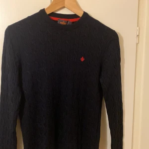 Mörkblå kabelstickad tröja Morris - Säljer en mörkblå kabelstickad tröja från Morris i 100% merinoull. Tröjan har ett rött broderat emblem på bröstet och rund hals. Långärmad och med ribbade muddar vid ärmslut och nederkant. Perfekt för dig som gillar klassisk stil.