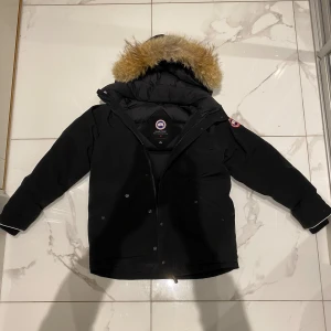 Svart dunjacka från Canada Goose - Säljer en svart dunjacka från Canada Goose med fluffig päls på huvan och klassisk logga på ärmen. Jackan har dragkedja och knappar framtill, två stora fickor och quiltad insida. Perfekt för kalla vinterdagar och riktigt snygg streetstil. 9/10 skick✅