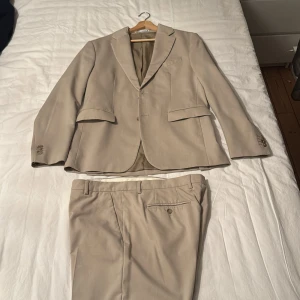 Beige kostym från Zara - Säljer en stilren beige kostym från Zara. Kavajen har klassisk passform med två fickor med lock och knappar vid ärmslut. Byxorna har raka ben, bälteshällor och bakficka med knapp. Perfekt för dig som vill ha en clean och modern look.
