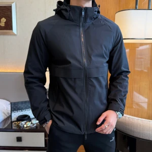 Svart vindjacka från Miu Miu - Svart vindjacka från Miu Miu med huva och hel dragkedja framtill. Jackan har diskret logga på bröstet och ärmen, samt praktiska fickor med dragkedja. Tillverkad i ett slätt syntetmaterial och har justerbara muddar med kardborre.