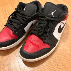 Nike Air Jordan 1 Low röd/svart/vit - Nike Air Jordan 1 Low sneakers i rött, svart och vitt med klassisk Jumpman-logga på plösen och Air Jordan-broderi på hälen. Skorna har snörning, rund tå och platt sula. Tillverkade i skinn med perforerade detaljer på tån för extra ventilation.