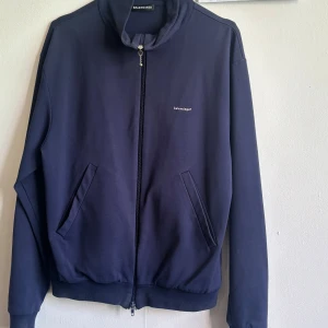 Mörkblå trackjacket från Balenciaga - Säljer en stilren mörkblå bomberjacka från Balenciaga med diskret logga på bröstet. Jackan har dragkedja framtill, två snedställda fickor och ribbade muddar vid ärmslut och nederkant. Perfekt för dig som gillar minimalistisk design och exklusiva märken.
