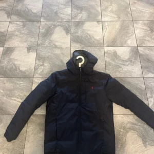 Mörkblå dunjacka från Polo Ralph Lauren - Snygg mörkblå dunjacka från Polo Ralph Lauren med huva och dragkedja framtill. Jackan har det klassiska röda Polo-logot broderat på bröstet och två sidofickor med dragkedja. Perfekt för kalla dagar och har en clean, sportig look. Litet märke som visas på tredje bilden, dock inget man tänker på 
