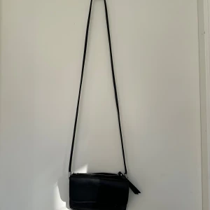 Svart axelväska i ”skinn” - En stilren svart axelväska i skinn med lång rem och dragkedja upptill. Väskan har ett minimalistiskt uttryck och insidan har flera fack för kort. Perfekt för dig som vill ha en enkel och snygg väska till vardags. Rymmer det viktigaste utan att bli klumpig.