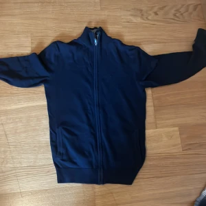 Mörkblå kofta från Pier One - Mörkblå kofta från Pier One med hel dragkedja framtill och ståkrage med grå insida. Tröjan har två fickor och är långärmad, perfekt för lager-på-lager. Enkel och stilren design som funkar till det mesta.