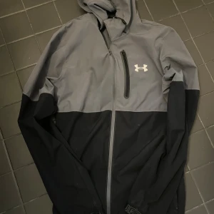 Grå och svart vindjacka Under Armour - Snygg tvåfärgad vindjacka från Under Armour i grått och svart. Jackan har huva, dragkedja framtill och en bröstficka med zip. Perfekt för dig som gillar sportig stil och vill ha något lätt och funktionellt. Logga på bröstet och detaljer på ärmslut.
