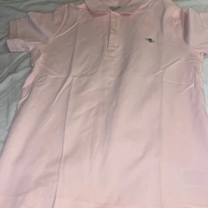 Ljusrosa pikétröja från GANT - Säljer en ljusrosa pikétröja från GANT med klassisk krage och knäppning framtill. Tröjan har korta ärmar och GANT:s broderade logga på bröstet. Tillverkad i mjuk bomull som känns skön mot huden. Perfekt för en fräsch och stilren look.