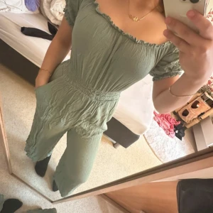 Grön jumpsuit från Amisu, strl 36 - Snygg ljusgrön jumpsuit från Amisu i storlek 36. Off-shoulder med volangkant och korta puffärmar. Byxorna är raka och har fickor. Materialet är mjukt och krinklat, perfekt för en chill och trendig look.